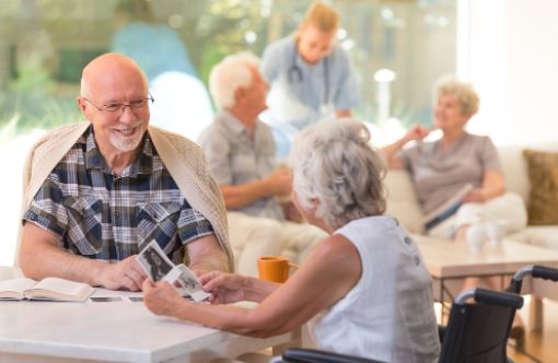 Elderly man and woman smiling and talking at a table while others chat in the background at a care home