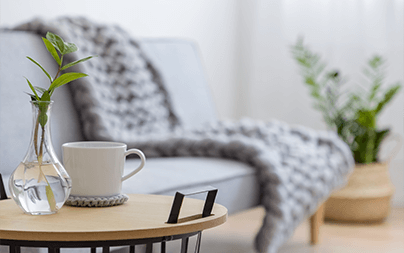 Contemporary living room with gray couch, cozy blanket, side table with plant and coffee cup.