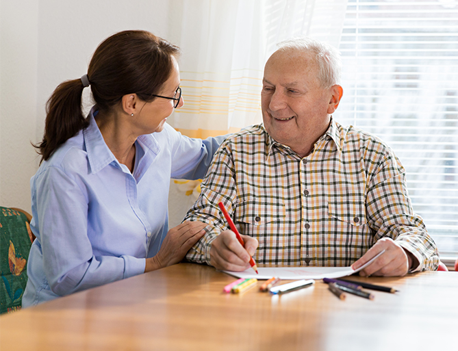 Elderly man coloring a page with red pencil, assisted by a smiling woman in a bright room