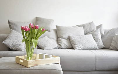 A comfortable grey sofa with several cushions and a wooden tray holding pink tulips and a teacup.