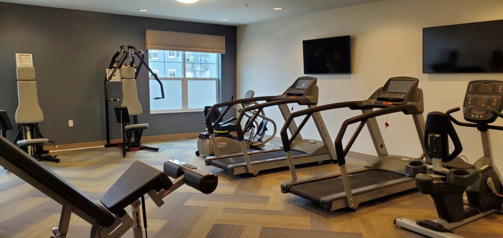 A well-lit fitness room with treadmills, exercise bikes, weight machines, and TV screens on the walls