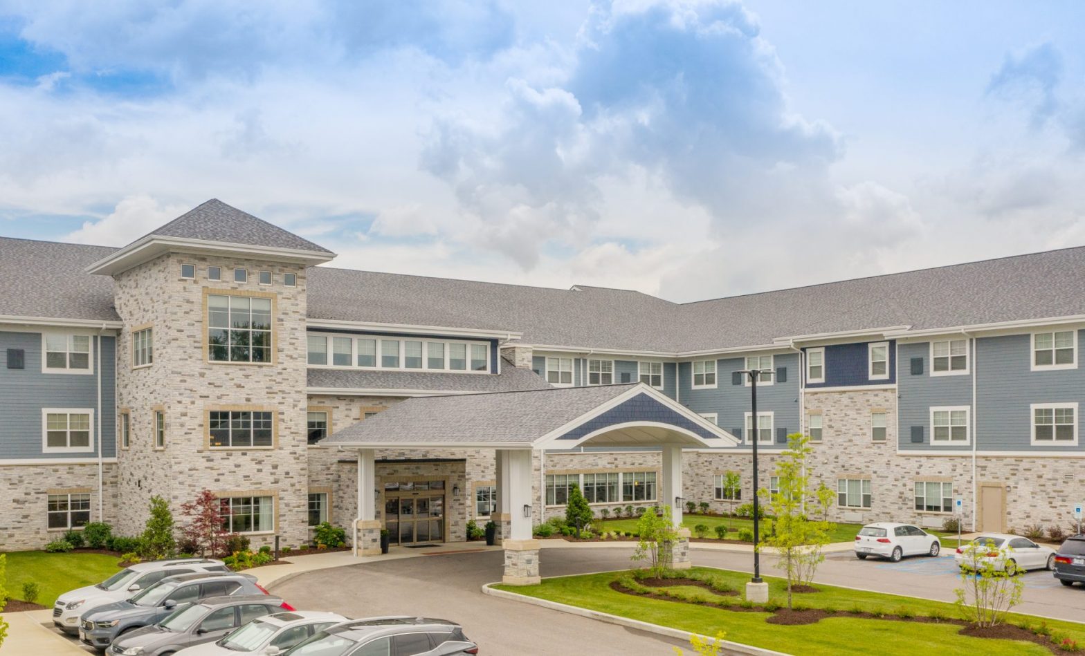 Modern senior living facility exterior with landscaped entrance and parked cars under a cloudy sky.
