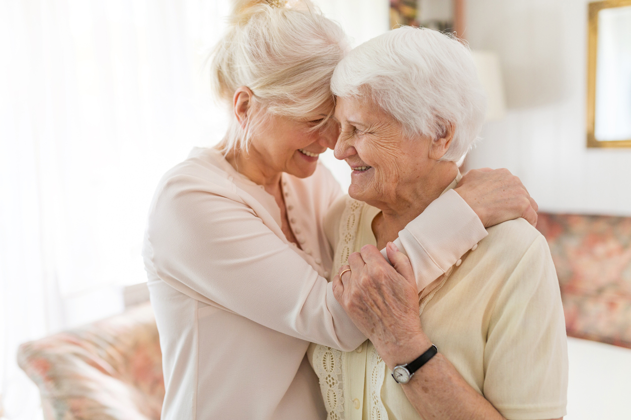 Two elderly women sharing a loving embrace and smiling warmly in a bright living room.