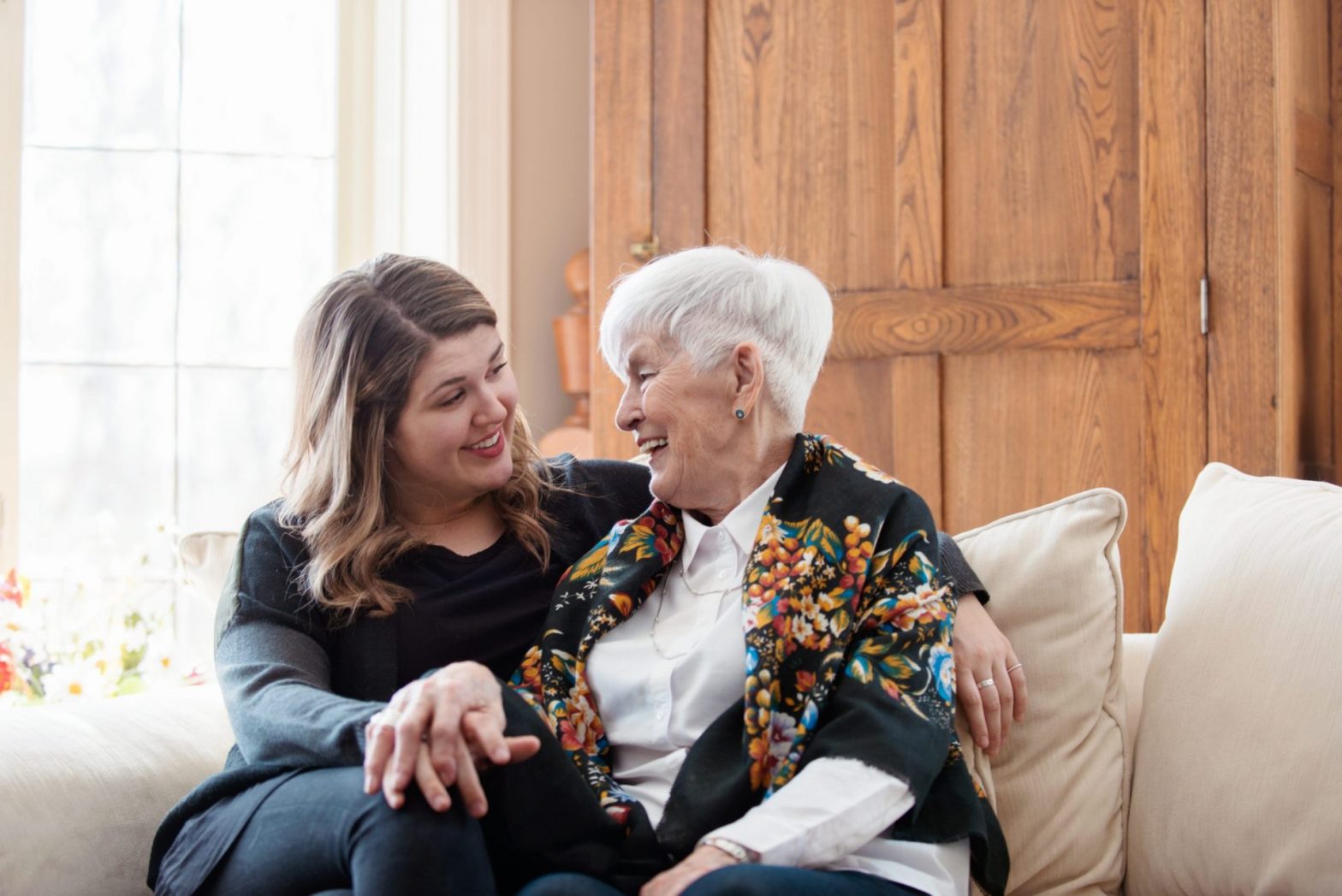Young woman and elderly woman smiling and embracing each other while sitting on a couch