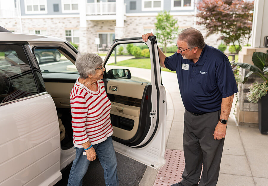 Senior woman assisted by staff member while exiting a white car at community entrance.