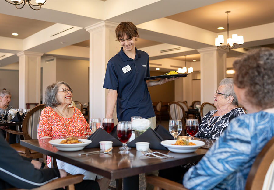 Residents enjoying a meal served by a staff member in a bright community dining room.