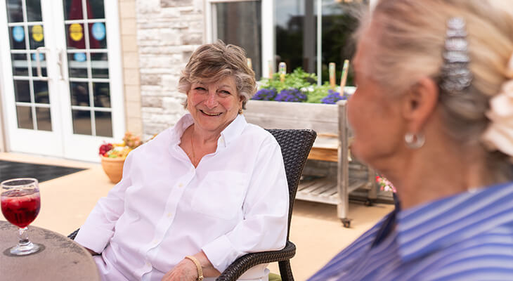 Two women conversing on a patio in a senior community, one has a drink beside her.