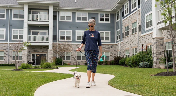 Elderly woman walking a dog on a path near a large residential building.