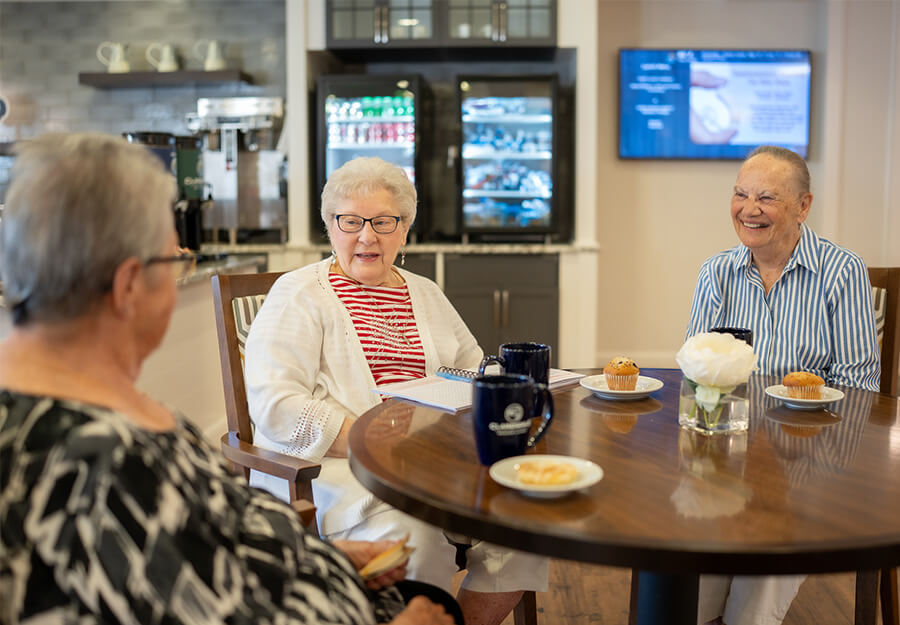Three seniors enjoy coffee and pastries at a community table in a common area.