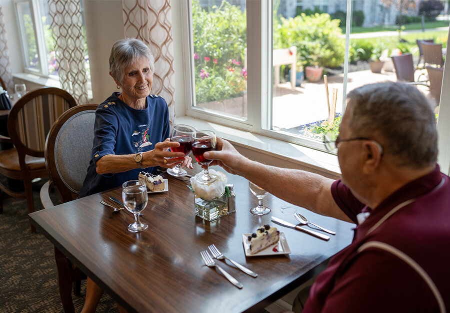 senior-living-dining-experience Two seniors enjoying dessert and drinks at a dining table near a large window.