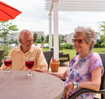 Two seniors enjoying drinks on a sunny community patio with garden view.